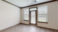 This single-floor residence offers a thoughtfully designed layout with a cohesive contemporary aesthetic throughout. The entryway opens directly adjacent to the kitchen, which features warm-toned shaker-style cabinetry, granite countertops, a subway tile backsplash, stainless steel appliances including a refrigerator, electric range, and built-in microwave, and a pendant light-adorned breakfast bar that connects seamlessly to the living area.

The living space is bright and open, benefiting from multiple windows flanking a private exterior door that provides views of surrounding greenery. Crown molding, a ceiling fan, and light wood-look luxury vinyl flooring contribute to a polished, cohesive feel. A dedicated dining or flex area mirrors this aesthetic, offering additional natural light.

The bedrooms are carpeted for comfort and include ceiling fans and ample natural light from window treatments. At least one bedroom features access to a walk-in closet equipped with built-in shelving and hanging racks. The bathroom is finished with granite countertops, warm wood cabinetry with brushed nickel hardware, a large framed mirror, and a full tub-shower combination with clean white surround.