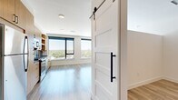 Located at 1300 40th Street in Denver's Cole neighborhood, this single-floor unit offers a thoughtfully designed open layout that combines the kitchen and living area into one cohesive, light-filled space. Light wood-tone luxury vinyl plank flooring runs throughout, complementing the clean white walls and creating a warm, contemporary feel.

The kitchen is a standout feature, outfitted with natural wood cabinetry, dark countertops, a blue-gray subway tile backsplash, and stainless steel appliances including a gas range, refrigerator, dishwasher, and microwave. A gold-tone faucet adds a refined accent. The living area is bright and airy, anchored by large floor-to-ceiling windows with black frames that frame sweeping views of Denver's treetops and skyline.

A hallway connects the main living space to the bathroom and closet, lined with paneled white barn doors featuring matte black hardware — a stylish nod to modern farmhouse design. The bathroom continues the unit's design language with a wood-tone vanity, dark countertop, matte black fixtures, a frameless glass walk-in shower with dark tile, and wood-look flooring. A ceiling fan and ample storage round out the functional and well-appointed interior.