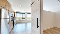 Located at 1300 40th Street in Denver's Cole neighborhood, this single-floor unit offers a thoughtfully designed open layout that combines the kitchen and living area into one cohesive, light-filled space. Light wood-tone luxury vinyl plank flooring runs throughout, complementing the clean white walls and creating a warm, contemporary feel.

The kitchen is a standout feature, outfitted with natural wood cabinetry, dark countertops, a blue-gray subway tile backsplash, and stainless steel appliances including a gas range, refrigerator, dishwasher, and microwave. A gold-tone faucet adds a refined accent. The living area is bright and airy, anchored by large floor-to-ceiling windows with black frames that frame sweeping views of Denver's treetops and skyline.

A hallway connects the main living space to the bathroom and closet, lined with paneled white barn doors featuring matte black hardware — a stylish nod to modern farmhouse design. The bathroom continues the unit's design language with a wood-tone vanity, dark countertop, matte black fixtures, a frameless glass walk-in shower with dark tile, and wood-look flooring. A ceiling fan and ample storage round out the functional and well-appointed interior.