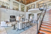 Located at 2370 Main St NW in Duluth, Georgia, this beautifully appointed space makes an immediate impression with its dramatic double-height ceilings and an open, airy layout that invites exploration. The main living area is anchored by light-toned herringbone hardwood flooring, which flows seamlessly throughout, adding a refined texture underfoot. Expansive floor-to-ceiling windows and glass doors flood the interior with abundant natural light while framing views of the outdoor amenity area, complete with a pool and courtyard beyond.

A grand staircase with sleek cable railings and a warm wood banister rises to an upper-level mezzanine overlooking the main floor, adding vertical drama and architectural interest. Elegant wainscoting and coffered panel walls in a crisp neutral palette lend a sophisticated, tailored character to every room.

The lounge area features a plush sectional sofa, accent chairs, and a sculptural low coffee table, creating a welcoming gathering space. Adjacent, a bar-height dining counter with upholstered swivel chairs accented by bold fabric patterns sits beneath softly lit arched niches showcasing curated artwork. The overall ambiance is one of polished, contemporary elegance—comfortable yet refined, with carefully considered details that elevate every corner of the space.