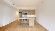 Located in Bethesda, MD, Unit 212 at 4909 Auburn Ave is a thoughtfully designed one-bedroom, two-bathroom residence offering a clean, modern aesthetic across a single floor. Light wood flooring runs throughout the space, complementing the bright white walls and creating a warm, cohesive atmosphere.

The open-concept kitchen is a standout feature, showcasing flat-front light wood cabinetry that extends to the ceiling, stainless steel appliances, a subtle tile backsplash, and a sleek white-topped island with matching wood accents. A linear pendant light anchors the kitchen, adding a contemporary touch. The living area flows naturally from the kitchen, bathed in natural light from a large window with outdoor views of surrounding greenery.

The primary bedroom offers a comfortable, well-proportioned retreat with two closets providing ample storage. Both bathrooms are finished with neutral tile flooring, wood-toned vanities with white quartz countertops, and large mirrors, reflecting the same modern sensibility found throughout the unit.

A hallway connects the private areas of the home, maintaining a logical and efficient layout. The overall ambiance is clean, minimalist, and move-in ready, making this Bethesda residence an excellent blend of style and functionality.