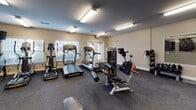 Located at 1001 Dunes St in Fredericksburg, Virginia, this fitness center offers a well-equipped, single-floor workout facility designed to support a wide range of exercise routines. The space is illuminated by bright overhead fluorescent lighting and features a neutral color palette with beige walls and durable carpet flooring throughout, creating a clean and functional environment.

The strength training area is generously outfitted with a full rack of dumbbells, kettlebells in varying weights, medicine balls, and multiple cable and weight machines, including a prominent multi-station cable system. Full-length mirrors line portions of the walls, enhancing the sense of space and allowing users to monitor their form during workouts. Flat weight benches are positioned throughout the floor for versatility.

The cardio section features treadmills, elliptical trainers, a stationary bike, and a stair climber, arranged to provide ample space between each machine. A mounted television is available for entertainment during cardio sessions. Natural light filters in through windows and glass-panel doors, adding a welcoming brightness to the cardio area.

Additional functional training equipment, including kettlebells and medicine balls neatly stored on dedicated racks, rounds out the facility's offerings. The layout is thoughtfully organized to maximize usable floor space while maintaining clear pathways between equipment zones.