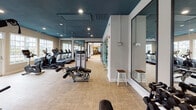 This fitness center at 4800 Waterford Point Dr in Raleigh, NC is a thoughtfully designed, single-floor facility offering a comprehensive workout experience across several distinct zones. The space is defined by a striking deep teal ceiling in the main workout area, contrasted by crisp white walls and warm neutral flooring, creating a modern and energizing atmosphere. Large windows line the perimeter, flooding the space with abundant natural light and providing views of the surrounding exterior. Full-length framed mirrors are strategically positioned throughout, enhancing the sense of openness and allowing users to monitor their form. The main floor features an extensive lineup of cardio equipment, including treadmills, ellipticals, and rowing machines, as well as a variety of strength training machines and free weight stations. A dedicated indoor cycling studio is enclosed by floor-to-ceiling glass panels, housing multiple spin bikes and a wall-mounted display screen for guided workouts. An additional functional training area includes a climbing wall and cable machines, catering to a wide range of fitness goals. Stylish industrial-style glass doors connect the fitness center to adjacent community spaces, reinforcing the open, integrated design of the overall facility.