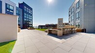 Located on the third floor of a modern Baltimore building, this expansive outdoor deck offers a thoughtfully designed amenity space with distinct zones for relaxation, socializing, and open-air leisure. The deck is finished with large-format porcelain pavers across its main terrace, creating a clean, contemporary aesthetic that complements the surrounding architecture's dark paneling and floor-to-ceiling glass facades.

Multiple areas of lush synthetic turf provide inviting green spaces, some bordered by glass railings that frame unobstructed views of the Baltimore waterfront and harbor beyond. Colorful lounge chairs and small side tables are arranged on these turf sections, ideal for taking in the sunlit skyline. A substantial built-in bar counter with a stone face and accompanying bar-height seating serves as a central social hub on the paved terrace.

Warm wood-clad raised planters filled with ornamental grasses add texture and visual interest throughout, while a pergola structure anchors one corner. Tiered wood steps lead between levels, and walkways connect the various zones seamlessly. The open-sky setting ensures abundant natural light throughout the day, making this deck a dynamic and versatile outdoor gathering space.