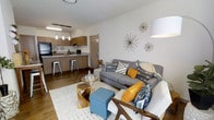 This single-floor space showcases a well-designed open-concept layout that seamlessly connects the kitchen and living area, creating a cohesive and functional environment. The kitchen features warm wood-toned cabinetry, a breakfast bar with industrial-style metal barstools, pendant lighting suspended from a track rail, and dark appliances. The countertops extend into a peninsula that serves as both a prep space and casual dining area, making the kitchen both practical and inviting.

The living area flows naturally from the kitchen, anchored by rich hardwood-style flooring throughout. A plush area rug defines the seating zone, which centers around a grey sofa accented with vibrant mustard yellow, teal, and patterned throw pillows, adding a lively, contemporary feel. A wood-accented coffee table and a teal upholstered ottoman provide functional yet stylish focal points. Decorative starburst wall art in a mid-century modern style adds a distinctive design touch to the neutral white walls.

The overall ambiance is warm, modern, and thoughtfully styled, balancing open connectivity with clearly defined functional zones. The consistent use of warm wood tones, neutral walls, and bold accent colors throughout gives the space a cohesive, curated aesthetic that is both comfortable and visually engaging.