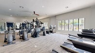 This well-appointed fitness center offers an expansive, single-floor layout designed to accommodate a full spectrum of workout needs. The space features a clean, contemporary aesthetic with neutral-toned walls, light carpet flooring, and recessed lighting complemented by ceiling fans that promote comfortable airflow throughout.

The cardio zone is generously outfitted with stationary bikes — including spin-style cycles with digital displays — treadmills, elliptical trainers, and stair climbers, all positioned to take advantage of the natural light streaming in through large, modern windows that frame outdoor views of surrounding greenery.

The strength training area is equally impressive, featuring a variety of cable and selectorized machines from brands such as Matrix, catering to both upper and lower body conditioning. A dedicated free weights section lines one wall, complete with a full rack of dumbbells, kettlebells, and additional strength accessories, all set in front of large floor-to-ceiling mirrors that enhance the sense of space and allow users to monitor their form.

Vaulted ceilings in portions of the facility add to the open, airy atmosphere, while the thoughtful layout ensures ample circulation space between equipment. The overall ambiance is bright, clean, and motivating — reflecting a professional-grade fitness environment built for everyday use.