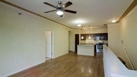 This single-story space showcases an open-concept layout that seamlessly connects the living area to the kitchen, creating a spacious and functional environment. The living area features warm hardwood flooring that extends throughout, complemented by crown molding along the ceiling perimeter and a ceiling fan with integrated lighting that enhances air circulation and illumination.

The kitchen is a standout feature, equipped with rich dark espresso cabinetry, granite countertops, and stainless steel appliances, including a gas range and built-in microwave. A peninsula with pendant lighting overhead provides additional counter space and serves as a natural dividing point between the kitchen and living area. Track lighting above the kitchen ensures ample task lighting, while a decorative wine rack adds a charming accent detail.

The neutral, light-toned walls create a bright and welcoming ambiance throughout. Multiple doorways visible from the main living space suggest additional rooms beyond, contributing to the overall functional flow of the layout. The combination of hardwood floors, modern finishes, and open design gives this space a polished yet comfortable character.