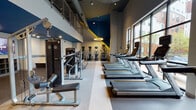 This fitness center in Ann Arbor, Michigan is a thoughtfully designed, full-featured facility that caters to a wide range of workout styles and fitness goals. The main workout floor is spacious and open, boasting soaring ceilings with exposed ductwork and striking circular halo pendant lights that lend an industrial-modern aesthetic. A bold color scheme of navy blue, gold, and gray carries throughout, accented by large geometric wall graphics that energize the space.

Floor-to-ceiling windows line one side of the main floor, flooding the area with natural light and offering views of the surrounding neighborhood — an ideal backdrop for cardio sessions on the row of Matrix treadmills positioned alongside them. Elliptical trainers and additional cardio equipment round out the aerobic offerings. The strength training section features Matrix cable machines, weight benches, free weights, and selectorized equipment, providing comprehensive options for resistance training.

A dedicated free weights and functional training room is adorned with a dramatic, full-wall motivational mural, creating an immersive and high-energy atmosphere. A separate cycling studio features stationary bikes arranged before a large display screen, complete with wall-mounted mirrors and resistance bands for supplementary training. The facility's durable, speckled flooring and efficient lighting throughout make it both practical and visually dynamic.