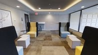 This single-floor space is designed as a contemporary collaborative lounge, offering a versatile environment ideal for informal meetings, group work, or social gatherings. The room features a tray ceiling with warm cove lighting that runs along the perimeter, complemented by recessed pot lights, creating a layered and inviting ambiance throughout.

The flooring consists of large-format carpet tiles in varying neutral tones — including beige, taupe, and brown — arranged in a patchwork pattern that adds visual texture to the space. The walls are finished in a light grey wood-effect panel, lending a clean, modern aesthetic.

Seating is modular and flexible, with high-back booth-style chairs in yellow and grey — each framed with dark privacy panels — providing semi-private nooks within the open layout. Accompanying white rectangular ottomans offer additional casual seating options. A large whiteboard or projection screen occupies one wall, supporting presentations or collaborative work sessions. A tall stool and a narrow countertop shelf are also present along the back wall, offering supplementary workspace. The overall design balances functionality with a welcoming, modern atmosphere.
