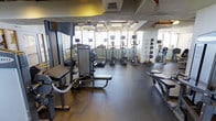 This fitness center offers a comprehensive workout environment designed to accommodate a variety of exercise routines. The space is outfitted with an impressive selection of equipment, including multiple treadmills, elliptical machines, and a stationary bike, all equipped with individual media screens for an engaging cardio experience. Strength training is well represented with a cable machine, weight bench, and additional resistance equipment positioned along one side of the floor. A row of medicine balls lines the wall, adding functional fitness options to the mix. The durable rubber flooring provides both safety and comfort underfoot throughout the workout area. Large windows along the far wall allow abundant natural light to fill the space, creating an open and energizing atmosphere. Full-length mirrors are strategically placed to assist with form and to visually expand the room. The exposed ceiling adds an industrial character to the space, while the overall layout is thoughtfully arranged to ensure ample room for movement between stations. This fitness center is a fully functional, well-maintained facility suited for a wide range of fitness levels and training styles.