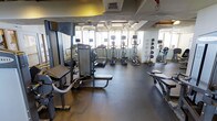 This fitness center offers a comprehensive workout environment designed to accommodate a variety of exercise routines. The space is outfitted with an impressive selection of equipment, including multiple treadmills, elliptical machines, and a stationary bike, all equipped with individual media screens for an engaging cardio experience. Strength training is well represented with a cable machine, weight bench, and additional resistance equipment positioned along one side of the floor. A row of medicine balls lines the wall, adding functional fitness options to the mix. The durable rubber flooring provides both safety and comfort underfoot throughout the workout area. Large windows along the far wall allow abundant natural light to fill the space, creating an open and energizing atmosphere. Full-length mirrors are strategically placed to assist with form and to visually expand the room. The exposed ceiling adds an industrial character to the space, while the overall layout is thoughtfully arranged to ensure ample room for movement between stations. This fitness center is a fully functional, well-maintained facility suited for a wide range of fitness levels and training styles.