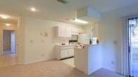This single-floor residence offers a clean, functional layout centered around an open-concept design that connects the kitchen and living areas seamlessly. The kitchen is outfitted with white cabinetry that extends to the upper walls, providing ample storage space. A built-in range, overhead exhaust hood, and a full-size refrigerator are among the key appliances featured. A peninsula counter serves as a natural divider between the kitchen and the adjacent living space, offering a convenient breakfast bar or serving area. The flooring transitions from tile in the kitchen to plush carpeting in the living area, adding a sense of warmth and comfort. Recessed and fluorescent lighting illuminate the space effectively, while a window near the kitchen allows for a degree of natural light. The neutral white and beige color palette throughout lends the space a bright, open feel that would serve as a versatile backdrop for a variety of interior styles. The overall layout is practical and well-organized, making efficient use of the available space.
