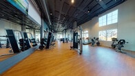 Located at 2525 Bayfront Blvd in Hercules, CA, this fitness center is a single-floor facility designed to support a comprehensive range of workout routines. The space features soaring ceilings with an exposed industrial-style framework, dark ceiling trusses, and ductwork that contrast with the warm hardwood flooring throughout, creating a modern yet welcoming athletic environment.

Natural light flows generously through a series of tall arched windows and large grid-pane windows along the perimeter walls, offering outdoor views of the surrounding landscape. The open floor plan is thoughtfully organized into distinct training zones. A dedicated cardio area lines one side of the facility, equipped with multiple treadmills and stationary bikes positioned to take advantage of the natural light and outdoor views. 

The strength training zone features a variety of cable machines and multi-functional weight stations, while tiered dumbbell racks provide a full range of free weights. A functional training alcove — accented by teal-painted walls — houses a cable functional trainer, a heavy bag, TRX suspension systems, and floor mats, ideal for dynamic workouts. Large wall mirrors are strategically placed throughout to maximize visibility and the sense of space. Track lighting and accent fixtures complete the well-lit, energetic atmosphere of this fitness facility.