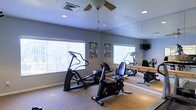 This single-floor fitness center offers a practical and functional workout space designed to meet everyday exercise needs. The room is outfitted with cardio equipment, including an elliptical trainer and a recumbent exercise bike, providing options for low-impact cardiovascular workouts. A full-length wall mirror spans one side of the room, visually expanding the space and allowing users to monitor their form during exercise. Fitness reference charts are displayed on the walls, offering guidance for proper technique and workout routines. Natural light filters into the room through multiple windows dressed with vertical blinds, creating a bright and energizing atmosphere. A ceiling fan with a light fixture is centrally mounted, promoting air circulation and comfort during workouts. Recessed lighting supplements the natural light, ensuring the space remains well-lit at all times. The flooring is finished with a durable, low-pile carpet suited to the demands of a fitness environment. A wall-mounted television is also visible in the mirror's reflection, adding an entertainment option for users during their sessions. The overall ambiance is clean, practical, and purposefully arranged to encourage regular physical activity.