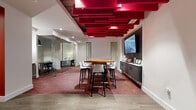 This thoughtfully designed single-floor office space makes an immediate visual impact through its striking suspended red ceiling installation — a series of layered, staggered panels that add dramatic depth and color to the main collaborative area. The ceiling detail anchors the open central zone, where a solid wood high-top table surrounded by contemporary bar-height chairs serves as a primary gathering point. A large wall-mounted display and a dark, built-in credenza complete this presentation-ready area, creating a functional hub for meetings and group discussions.

The patterned red and grey carpeting throughout reinforces the bold color palette while defining the transition between meeting zones and circulation areas. Wide-plank hardwood flooring at the entry provides a warm contrast to the carpeted interior.

Glass-walled partitions divide the space into several enclosed meeting rooms, each furnished with round tables, pendant lighting, and dedicated display screens — offering privacy without sacrificing visual openness. A utility area with dark tile backsplash, cabinetry, and workspace equipment supports day-to-day office needs. A boldly painted red accent wall adds further energy to the support corridor. Together, the design balances collaborative flexibility with private, focused workspaces in a cohesive and professional environment.