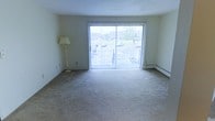This single-floor residential space is designed with functionality and comfort in mind. The living area serves as the focal point of the unit, featuring large sliding glass doors that span nearly the full width of the room, allowing an abundance of natural light to fill the interior. The doors open to a balcony, offering views of the surrounding outdoor environment, including greenery and a parking area beyond. The room is finished with neutral-toned wall-to-wall carpeting, providing a soft and cohesive feel underfoot. Light-colored walls contribute to the open and airy atmosphere of the space. A baseboard heating system runs along the exterior wall beneath the sliding doors, ensuring efficient and consistent warmth throughout the colder months. Vertical blinds on the sliding doors offer privacy and light control as needed. The overall layout is open and unobstructed, allowing for flexible furniture arrangement to suit a variety of living styles. The neutral color palette and ample natural light create a calm and welcoming ambiance throughout the space.