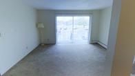 This single-floor residential space is designed with functionality and comfort in mind. The living area serves as the focal point of the unit, featuring large sliding glass doors that span nearly the full width of the room, allowing an abundance of natural light to fill the interior. The doors open to a balcony, offering views of the surrounding outdoor environment, including greenery and a parking area beyond. The room is finished with neutral-toned wall-to-wall carpeting, providing a soft and cohesive feel underfoot. Light-colored walls contribute to the open and airy atmosphere of the space. A baseboard heating system runs along the exterior wall beneath the sliding doors, ensuring efficient and consistent warmth throughout the colder months. Vertical blinds on the sliding doors offer privacy and light control as needed. The overall layout is open and unobstructed, allowing for flexible furniture arrangement to suit a variety of living styles. The neutral color palette and ample natural light create a calm and welcoming ambiance throughout the space.