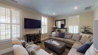 Located in Columbus, Georgia, this single-floor space offers a warm and inviting atmosphere with thoughtfully curated design details throughout. The main lounge area is anchored by a generously sized sectional sofa and coordinating accent chairs, all arranged around a tufted ottoman that serves as a central gathering point. A large wall-mounted flat-screen television is positioned above a dark wood console table with decorative carved detailing, flanked by windows fitted with classic white plantation shutters that allow soft, natural light to filter through.

Crown molding lines the ceiling throughout the space, lending a refined architectural touch. Recessed lighting complements the natural light, maintaining a bright yet comfortable ambiance. Framed artwork adorns the walls, adding visual interest and a curated feel to the room.

A standout feature is the elegant French door with glass pane insets, which separates the main lounge from an adjoining alcove. This secondary area includes a built-in kitchenette with a dark granite countertop, gray cabinetry, a stainless steel refrigerator, and open upper shelving stocked with books — ideal for a reading nook or refreshment station. The neutral color palette of warm beige tones and gray upholstery ties the entire space together cohesively.