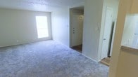 This single-floor space presents a straightforward, functional layout centered around a carpeted main living area finished in a neutral gray tone. The room is illuminated by a window along one wall, allowing natural light to brighten the interior and complement the light-colored walls throughout.

A defined entryway separates the main living area from the front door, featuring hard surface flooring that transitions cleanly into the carpeted space. The front door is solid and paneled, providing a clear boundary between the entry corridor and the living area. A textured popcorn ceiling runs throughout, adding a classic residential character to the space.

The walls are painted in a consistent light gray-white tone, creating a neutral backdrop that suits a variety of furnishing styles. Electrical outlets and light switches are positioned at standard heights along the walls. A secondary door is visible along the right wall, suggesting access to an additional room or closet area.

Overall, the space maintains a clean, unfurnished condition, offering a blank canvas for personalization. The combination of carpeted floors, natural light, and a defined entryway contributes to a practical and accessible layout.