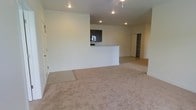 This single-floor space is designed with an open-concept layout that connects the living area to the kitchen, creating a seamless flow throughout. The living area features wall-to-wall carpeting in a neutral beige tone, providing a comfortable and cohesive base for the space. Recessed lighting is installed throughout the ceiling, offering consistent, clean illumination across the unit.

The kitchen is positioned toward the rear of the open area and is defined by a raised breakfast bar that serves as a natural divider between the cooking and living spaces. Dark-toned cabinetry lines the kitchen walls, providing a subtle contrast against the light-colored walls and countertops. A microwave is visible among the kitchen appliances, hinting at a functional setup.

Adjacent to the kitchen, a doorway leads to what appears to be a walk-in closet or dedicated storage room with built-in shelving, offering practical organizational space. The walls throughout are finished in a clean, neutral white, contributing to a bright and open atmosphere. The overall design is straightforward and functional, maximizing usable space within the single-floor footprint.