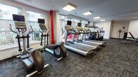 Located at 1810 Highpoint Ave in Richmond, Virginia, this well-equipped fitness center offers a comprehensive workout environment across a single floor. The space is thoughtfully designed with distinct zones to accommodate a full range of exercise needs.

The cardio area is lined with large windows that flood the space with natural light and provide views of the surrounding community, creating an energizing atmosphere for workouts. A row of Life Fitness treadmills and elliptical trainers are positioned to take advantage of these outdoor views, while a stair climber rounds out the cardio offerings.

The strength training section features a Synergy360 functional training system alongside cable machines, a rowing machine, stability balls, and medicine balls, all complemented by full-length mirrors that enhance the sense of space. Adjustable benches and a dumbbell rack provide additional free-weight options in a dedicated corner of the facility.

Bold red accent walls in portions of the space add a dynamic, high-energy aesthetic that contrasts with the clean white walls and dark patterned carpet flooring found throughout. Recessed panel lighting ensures consistent illumination across all workout zones. A sanitation station with wipes and hand sanitizer is conveniently located near the entrance, reflecting a commitment to cleanliness and user comfort. Overall, the fitness center delivers a modern, functional, and inviting environment for residents.
