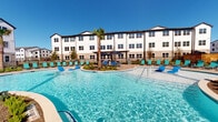 Located at 19770 Clay Rd in Katy, Texas, this outdoor amenity space is a standout feature of a modern residential community, thoughtfully designed to encourage relaxation and social engagement. At the heart of the space is a generously sized resort-style swimming pool with crystal-clear water, surrounded by teal lounge chairs and a central pergola structure shaded by a towering palm tree — creating a vibrant, vacation-inspired atmosphere under the Texas sky.

The covered patio area is anchored by sturdy natural stone columns and finished with rich, multi-toned paver flooring that adds warmth and texture to the environment. A ceiling fan overhead enhances comfort during warmer months, while a white dining table and chairs offer an inviting outdoor dining setting. Large black-framed windows provide a visual connection between the interior clubhouse and the outdoor pool deck, seamlessly blending indoor and outdoor living.

Adjacent lounge areas feature plush outdoor seating nestled beneath the covered walkway, with views of a manicured lawn and gated recreational green space. The surrounding multi-story buildings are clad in white horizontal siding with bold dark trim, contributing to a clean, contemporary coastal aesthetic. Lush tropical landscaping, including palm trees and native plantings, completes the polished, resort-like character of this community amenity space.