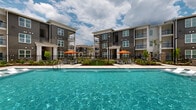 This expansive outdoor pool amenity in Scott, Louisiana offers a well-appointed resort-style retreat designed for relaxation and recreation. At the heart of the space is a generously sized main swimming pool with crystal-clear turquoise water, complemented by a separate, connected spa pool — ideal for unwinding after a long day. The pool deck is finished in light-toned pavers and features an abundance of chaise lounges and seating arrangements to accommodate a variety of guests.

Vibrant orange umbrella-shaded seating clusters are thoughtfully distributed throughout the deck, providing sun protection alongside an eye-catching pop of color. A covered outdoor lounge area anchors one side of the space, outfitted with comfortable navy blue upholstered furniture, a ceiling fan, and a modern pergola with warm wood-toned beams — creating a shaded social gathering spot. An additional pergola structure with a slatted wood roof offers a more intimate shaded seating area near the pool's edge.

The entire pool area is framed by manicured landscaping, including neatly trimmed greenery and planted garden beds, with iron fencing providing a clean boundary. The backdrop consists of contemporary three-story residential buildings featuring dark gray siding, private balconies, and large windows, lending the space a polished, modern community aesthetic beneath the open Louisiana sky.