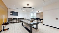 Located at 1600 James Naismith Dr in Ottawa, Ontario, this contemporary games room — branded "Level Up Your Game" — is a thoughtfully designed recreational space that offers a diverse range of entertainment options. The room features light hardwood-style flooring throughout and a crisp white ceiling accented by a bold circular ring pendant light that serves as a striking focal point overhead.

A full-size pool table anchors the centre of the room, complemented by a shuffleboard table, a foosball table, and a stylish round poker table surrounded by sleek leather chairs. A built-in bar-height counter with stools lines one wall beneath a wall-mounted flat-screen television, providing a comfortable spectator area. Open shelving nearby offers additional storage and display space.

The design blends energetic amber-orange accent walls with clean neutral tones and natural wood slat panelling near the branded signage wall, creating a dynamic yet polished atmosphere. Large floor-to-ceiling windows flood a dedicated seating corner with natural light and provide views of the surrounding outdoor environment. A frosted-glass entry door adds a refined touch at the entrance. The overall ambiance is modern, social, and inviting — purpose-built for leisure and friendly competition.