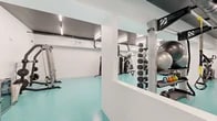 This expansive fitness center offers a comprehensive workout environment across a single, open floor plan. The space is designed with a clean, modern aesthetic, featuring crisp white walls, a striking teal rubberized flooring surface, and bright linear fluorescent lighting that ensures excellent visibility throughout. Exposed ductwork along the ceiling adds an industrial character to the otherwise polished interior.

The facility is organized into distinct functional zones. A strength training area houses a multi-station cable machine, a full dumbbell rack, weight plates, kettlebells, and stability balls, all arranged for ease of access and safe movement. Full-length wall mirrors in this section allow users to monitor their form during exercises. A dedicated cardio zone features a row of spin bikes positioned in front of a wall-mounted television screen, accompanied by additional mirrors and a step platform area suitable for aerobic classes. Larger cardio machines, including an elliptical trainer and rowing-style equipment, occupy a separate open section of the floor.

The space benefits from split-system air conditioning units mounted along the walls, ensuring a comfortable training environment. The open column structure helps define different workout areas while maintaining a spacious, uncluttered feel. The overall ambiance is clean, functional, and purpose-built for a wide range of fitness activities.