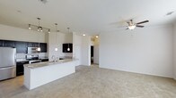 This single-floor residence in Frisco offers a thoughtfully designed open-concept layout that seamlessly connects the kitchen and living areas. The kitchen is appointed with rich dark cabinetry, granite countertops, a stainless steel sink, and stylish pendant lighting that hangs above the breakfast bar peninsula — providing both functional workspace and casual seating. Track lighting adds additional illumination to the kitchen zone, ensuring a bright and practical cooking environment. The open living area flows naturally from the kitchen, featuring plush wall-to-wall carpeting underfoot and a ceiling fan overhead that contributes to the room's comfortable, airy atmosphere. White walls throughout the space keep the interior feeling clean and bright. A central hallway leads to additional rooms, including what appears to be a bedroom and bathroom, each accessed through classic panel doors. The neutral color palette and well-considered layout make the space highly adaptable to a variety of furniture arrangements and personal styles. Overall, this Frisco residence delivers a welcoming, functional environment suited for everyday living.