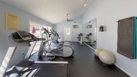 This fitness center is housed in a single-story space designed to support a comprehensive workout experience. The room features vaulted ceilings that lend an open, airy feel, complemented by light blue walls and a dark rubberized floor that is both functional and easy to maintain. Ceiling fans are positioned throughout to promote air circulation during workouts.

The cardio section is well-stocked with multiple Precor elliptical trainers, treadmills, and upright and recumbent stationary bikes, arranged in an organized row to maximize floor space. A multi-station cable weight machine anchors the strength training area, accompanied by a flat bench and stability balls for functional fitness exercises. Track lighting along one wall provides targeted illumination across the equipment.

Full-length mirrors line one side of the room, visually expanding the space while allowing users to monitor their form. Large windows and a glass-panel door along the opposite wall flood the interior with natural light and offer views of what appears to be an outdoor pool and patio area. The overall ambiance is clean, practical, and welcoming, making it a functional facility for a variety of fitness routines.
