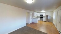 This single-floor residence offers a thoughtfully designed open-concept layout that seamlessly connects the living and kitchen areas. The kitchen is a functional and modern space, featuring crisp white cabinetry, a stainless steel refrigerator, electric range, and built-in microwave. A peninsula with a dark countertop provides additional workspace and serves as a natural divider between the kitchen and the adjoining living area. Light wood-look flooring runs throughout the kitchen, transitioning into soft carpeting in the living space, adding warmth and visual distinction between the two zones. The interior is finished with clean white walls and white trim, lending a bright and neutral backdrop that enhances the sense of openness. Flush-mount ceiling lighting provides consistent illumination throughout. A doorway visible from the living area suggests access to additional rooms or storage, including what appears to be a closet with slatted doors. The overall ambiance is tidy, functional, and move-in ready, appealing to those who value a clean, contemporary aesthetic with practical, everyday convenience built into the design.