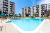 This residential complex is composed of multiple mid-rise brick buildings, each rising several stories with private balconies offering elevated outdoor space for residents. The buildings are arranged to frame a central outdoor amenity area, which serves as the social hub of the property.

The outdoor area centers on a generously sized swimming pool with clear, turquoise water, surrounded by a spacious concrete deck furnished with lounge chairs and teal patio umbrellas. A children's playground is situated nearby, making the space suitable for a range of residents. Landscaping with mature trees and shrubs adds greenery throughout the common grounds.

The building's lobby entrance features a utilitarian double-door entry with narrow sidelights, metal kick plates, and tile flooring. The interior corridor is finished with warm cream-toned walls accented by layered white chair rail and wainscoting molding, lending a classic, structured aesthetic to the common hallways. Wall-mounted sconces provide soft ambient lighting, and standard safety features including fire alarm components and exit signage are prominently placed throughout. The overall ambiance of the shared interior spaces is functional and well-maintained, reflecting a straightforward residential design with traditional architectural detailing.