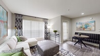 This single-floor residence offers a thoughtfully designed layout that balances functionality with contemporary style. The living area serves as the heart of the unit, featuring wood-style plank flooring, neutral grey walls, and large windows dressed with horizontal blinds and patterned curtains that allow generous natural light to fill the space. A dedicated dining nook with a round table sits adjacent to the living area, reinforcing an open, connected feel. The entry door, framed by a sidelite window with plantation shutters, contributes additional brightness to the common areas.

The kitchen is well-equipped with dark espresso cabinetry, light granite-style countertops, a full suite of black appliances including a refrigerator, electric range with overhead microwave, and dishwasher. A peninsula counter provides a convenient transition between the kitchen and the living space.

The unit includes two bedrooms. One bedroom features plush teal carpeting, a bold accent wall, and a large window with sheer curtains, while the second offers wood-style flooring, a ceiling fan, and natural light. The bathroom is finished with dark cabinetry, granite-style countertops, a large mirror, and a tub-shower combination with white tile surround.
