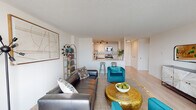 This thoughtfully designed single-floor residence offers a seamless blend of modern style and functional living. The open-concept living and dining area is anchored by large floor-to-ceiling windows that flood the space with natural light and frame expansive city views. Light hardwood flooring runs throughout the main living areas, complementing the bright white walls and creating a clean, contemporary aesthetic.

The living room comfortably accommodates a full seating arrangement and flows naturally into a designated dining space positioned near the window wall, providing a pleasant backdrop for everyday meals. A decorative credenza and arched gold shelving unit add character to the room.

The pass-through kitchen features white cabinetry with matte black hardware, a checkered tile backsplash, light quartz countertops, a deep single-basin sink, and stainless steel appliances. A breakfast bar with seating connects the kitchen to the main living area, enhancing its open feel.

The bedroom is a serene retreat with plush carpeting, a generous built-in closet, and a large window offering city views. The bathroom is clean and functional, finished with white tile and a full shower-tub combination. Overall, the residence offers a cohesive, well-lit living environment with a modern, eclectic design sensibility.