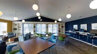 This single-floor space is designed as a modern communal area, combining functionality with a welcoming aesthetic. The room's most striking architectural feature is its vaulted ceiling, which creates an expansive, airy atmosphere enhanced by a series of spherical pendant lights that provide warm, even illumination throughout.

The space is furnished with multiple dark-wood rectangular tables paired with a mix of vibrant green and teal modern chairs, creating a flexible layout well-suited for group work, study, or casual gatherings. A built-in shelving unit lines one wall, offering display and storage space, while a workstation with a computer terminal is conveniently positioned nearby, suggesting a business or resource center function.

Bold navy blue accent walls contrast sharply with the white vaulted ceiling, lending the space a contemporary feel. Large windows and glass doors along one wall flood the interior with natural light and frame views of lush, snow-dusted trees outside. A lounge area with upholstered seating is visible to one side, adding a relaxed counterpoint to the workspace tables. The patterned carpet anchors the room's design scheme cohesively.
