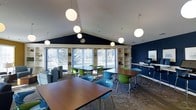 This single-floor space is designed as a modern communal area, combining functionality with a welcoming aesthetic. The room's most striking architectural feature is its vaulted ceiling, which creates an expansive, airy atmosphere enhanced by a series of spherical pendant lights that provide warm, even illumination throughout.

The space is furnished with multiple dark-wood rectangular tables paired with a mix of vibrant green and teal modern chairs, creating a flexible layout well-suited for group work, study, or casual gatherings. A built-in shelving unit lines one wall, offering display and storage space, while a workstation with a computer terminal is conveniently positioned nearby, suggesting a business or resource center function.

Bold navy blue accent walls contrast sharply with the white vaulted ceiling, lending the space a contemporary feel. Large windows and glass doors along one wall flood the interior with natural light and frame views of lush, snow-dusted trees outside. A lounge area with upholstered seating is visible to one side, adding a relaxed counterpoint to the workspace tables. The patterned carpet anchors the room's design scheme cohesively.