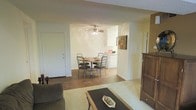 This single-floor space offers an open and functional layout that connects the main living areas in a cohesive, easy-to-navigate flow. The living room provides a comfortable gathering space with soft carpeting and room for seating, anchored by a low-profile coffee table. The space opens directly into a dedicated dining area, which features warm hardwood-style flooring, a round dining table with seating for four, and a ceiling fan overhead that adds both style and ventilation. Natural light fills the dining area, creating a bright and welcoming atmosphere. Just beyond, the kitchen is efficiently arranged with white cabinetry, appliances, and ample counter space, making it practical for everyday meal preparation. A storage unit or hutch near the living area provides additional functional storage. The neutral color palette throughout — consisting of soft whites and warm wood tones — lends a clean, understated aesthetic. The open-concept design between the living, dining, and kitchen zones allows for versatile use of the space, making it ideal for both quiet daily routines and casual entertaining.
