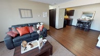 This single-floor unit offers an open and functional layout that seamlessly connects the living area to the kitchen and dining space. The living room is a welcoming area finished with plush carpet underfoot and neutral-toned walls that create a warm, inviting atmosphere. A textured popcorn ceiling adds a classic touch overhead, while large windows along one wall allow natural light to filter into the space, enhancing the bright and airy feel. A wall-mounted air conditioning unit and baseboard heater provide year-round climate control.

Transitioning from the living room, the flooring shifts to a rich hardwood-style surface that extends through the open-concept area toward the kitchen. The kitchen features dark cabinetry that contrasts attractively with the lighter walls, providing ample storage space. The layout supports easy flow between the kitchen and dining area, making the space practical for everyday use.

A dark interior door visible from the living area leads to additional private spaces within the unit. Overall, the space balances comfort and practicality with a cohesive, neutral color palette accented by warm wood tones throughout.