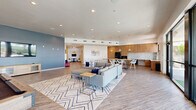 Located at 3510 N Craycroft Rd in Tucson, Arizona, this generously proportioned single-floor community space offers a modern, open-concept design ideal for gathering and recreation. The layout flows seamlessly from zone to zone, unified by durable wood-look flooring and a clean, contemporary aesthetic throughout.

The kitchen area is a standout feature, outfitted with warm wood cabinetry extending to the ceiling, a marble-style tile backsplash, black stainless steel appliances including a full-size refrigerator, built-in oven, and a large communal dining table with stylish wire chairs, illuminated by modern pendant lighting above.

The main lounge area is anchored by a large sectional sofa set atop a geometric-patterned area rug, complemented by a pool table nearby. Floor-to-ceiling windows and glass doors flood the space with abundant natural light and open to an outdoor terrace with sweeping views of the surrounding Tucson landscape.

A dedicated game nook features bold blue diagonal stripe accent walls, a wall-mounted flat-screen TV, and a round game table with seating. Additional accent walls in muted slate blue, a decorative peace sign, textured stone wall panels, and recessed lighting throughout contribute to the space's inviting, vibrant ambiance.