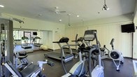 This fitness center offers a thoughtfully organized workout space spread across a single floor. The room is outfitted with a range of cardio equipment, including treadmills and elliptical trainers, positioned to allow comfortable use and easy circulation between machines. A stability ball and additional free weights are also available, catering to varied fitness routines. The flooring is a durable dark rubber material, well-suited for high-traffic exercise environments. Full-length mirrored walls line one side of the room, visually expanding the space and allowing users to monitor their form during workouts. The ceiling features recessed lighting alongside a ceiling fan, ensuring the room remains well-lit and properly ventilated. Track lighting adds supplemental illumination, while vertical blinds along the walls offer control over natural light. The overall ambiance is clean, functional, and practical, with a neutral color palette of whites and light greens that contributes to a bright and focused atmosphere. The layout maximizes usable floor space while maintaining an open, accessible feel suited for individual fitness activity.