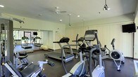 This fitness center offers a thoughtfully organized workout space spread across a single floor. The room is outfitted with a range of cardio equipment, including treadmills and elliptical trainers, positioned to allow comfortable use and easy circulation between machines. A stability ball and additional free weights are also available, catering to varied fitness routines. The flooring is a durable dark rubber material, well-suited for high-traffic exercise environments. Full-length mirrored walls line one side of the room, visually expanding the space and allowing users to monitor their form during workouts. The ceiling features recessed lighting alongside a ceiling fan, ensuring the room remains well-lit and properly ventilated. Track lighting adds supplemental illumination, while vertical blinds along the walls offer control over natural light. The overall ambiance is clean, functional, and practical, with a neutral color palette of whites and light greens that contributes to a bright and focused atmosphere. The layout maximizes usable floor space while maintaining an open, accessible feel suited for individual fitness activity.