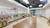 Located at 4710 Bethesda Ave in Bethesda, Maryland, this single-floor commercial space offers a functional and adaptable layout well-suited for a variety of business uses. The space features an open floor plan with light tile flooring throughout most areas, transitioning to wood-tone flooring in one section. Ceilings are finished with a dark painted, open-plenum design that exposes ductwork, wiring conduits, and ceiling fans alongside flush-mounted fluorescent light panels, lending an industrial character to the interior. The main entry area is bright and welcoming, with large frosted-glass storefront windows and doors that allow natural light to filter in while maintaining privacy. Views of mature greenery are visible through the upper clerestory windows. The primary open area provides generous floor space with wire metro shelving units on casters, a central work table, and ample room for storage or display. A secondary open room features additional shelving, a clothes rack, and movable partition screens, and connects to a back door access point. Exposed HVAC systems and utility infrastructure are visible throughout, indicating the space is in a transitional or build-out phase. The overall layout is practical and highly configurable, making it a strong candidate for retail, a showroom, or a service-oriented commercial operation in the heart of Bethesda.