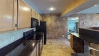 This single-floor space offers a thoughtfully designed interior with a striking blend of textures and finishes throughout. The kitchen is a functional and stylish centerpiece, featuring light maple cabinetry paired with rich, dark granite countertops. Black appliances — including an electric range, built-in microwave, and refrigerator — provide a sleek contrast to the warm cabinetry tones. Tile flooring ties the space together, while an eye-catching faux-finish accent wall in deep earthy browns and golds adds a dramatic, artistic touch.

The living area continues the bold design aesthetic, with artisan-applied faux plaster walls rendered in warm taupes and muted earth tones that lend a sophisticated, textured ambiance. Recessed lighting illuminates the space from above, complementing the plush carpeted flooring underfoot. A built-in ledge runs along one wall, offering both architectural detail and functional display space. The seating arrangement, anchored by a curved leather sofa and matching accent chair in rich caramel tones, creates a comfortable and inviting atmosphere.

The 3D scan further reveals a dramatic great room with soaring double-height ceilings, floor-to-ceiling windows that flood the space with natural light, and an elegant staircase — adding grandeur to the overall layout.
