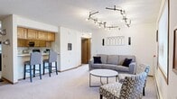 This single-floor unit offers a thoughtfully designed layout that balances comfort and functionality. The open-concept living area is a standout feature, anchored by a striking circular track lighting fixture that adds a modern, decorative element to the space. Plush carpeting runs throughout the living room, which accommodates a sofa, accent chairs, and a round coffee table, while a window provides natural light and an outdoor view. The living area flows seamlessly into the kitchen, which is separated by a breakfast bar with bar seating. The kitchen is well-equipped with warm oak cabinetry, black appliances including a refrigerator, electric range, and dishwasher, and tile flooring. An adjacent dining nook with a ceiling fan offers a defined space for meals. The bedroom is generously sized, featuring soft carpet, a neutral accent wall in a muted grey tone, a window with natural light, and direct access to a private bathroom. That bathroom includes a walk-in shower, tile flooring, a wood-trimmed vanity, and a large mirror. A second bathroom, also featuring a wood vanity and large mirror, serves the remainder of the unit. Throughout the space, warm oak door and trim details provide a consistent and cohesive aesthetic.