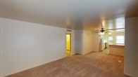 This single-floor residence offers a straightforward and functional layout designed for comfortable everyday living. The main living area is spacious and open, featuring neutral wall tones and wall-to-wall carpeting in a warm beige hue that adds softness underfoot throughout the space. A ceiling fan is centrally mounted, providing both airflow and a subtle decorative touch to the room.

The open floor plan allows the living area to flow naturally toward the kitchen, which is visible in the background and features light cabinetry and standard appliances, including a refrigerator. The kitchen benefits from its own lighting, creating a warm, well-lit workspace. A doorway off the main living area leads to an additional room, glimpsed with warm lighting and matching carpeting, suggesting a bedroom or utility space.

The smooth, painted ceilings and clean wall finishes throughout give the space a tidy, move-in-ready appearance. Ventilation is provided via ceiling-integrated vents. The overall ambiance is clean, neutral, and practical, making it a versatile space that can be easily personalized. The layout prioritizes an efficient use of space, with clear sightlines from the living area through to the kitchen and adjoining rooms.