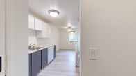 This single-floor unit offers a clean, neutral aesthetic carried consistently throughout its layout. Light wood-look plank flooring extends across all rooms, providing a cohesive and modern foundation. The galley-style kitchen is efficiently designed with a two-tone cabinet scheme — white upper cabinets paired with slate-blue lower cabinets — complemented by light countertops, a double sink, a dishwasher, a refrigerator, and an oven. Ceiling-mounted flush lighting illuminates the kitchen, which opens toward the main living areas.

The living room serves as a standout space, anchored by a brick fireplace with a classic surround that adds character and warmth. A ceiling fan with a light fixture provides air circulation, and large vertical-blind-covered sliding doors allow natural light to fill the room. Adjacent rooms include multiple bedrooms featuring windows with views of mature, leafy trees, white walls, and generous closet space. The bedrooms maintain the same light flooring and bright, neutral palette found throughout. Overall, the unit presents a well-maintained, move-in-ready environment with a clean and functional design.