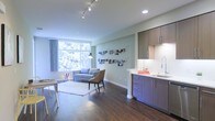 This single-floor open-concept space combines living and kitchen areas into a cohesive, efficiently designed layout. The rich dark hardwood flooring anchors the interior, providing a warm contrast to the soft, neutral-toned walls painted in a calming light grey-green hue. A large floor-to-ceiling window on the far wall floods the living area with abundant natural light and frames a verdant, tree-filled outdoor view, creating a strong connection to the surrounding environment. The living area accommodates a grey sofa, accent chair, and a casual work desk, all positioned to take advantage of the natural light. A plush white area rug defines the seating zone atop the hardwood floor. Track lighting along the ceiling supplements natural light throughout the space. The kitchenette, neatly integrated along one wall, features dark wood-toned cabinetry both above and below the counter, a white countertop with a deep undermount sink, and a modern gooseneck faucet. Recessed lighting above the kitchen ensures ample task illumination. The overall ambiance is clean, contemporary, and functional, with a thoughtful blend of materials and finishes that create an inviting, modern atmosphere.