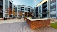 This expansive courtyard amenity space is thoughtfully designed to serve as a central hub for outdoor living and recreation within a modern residential community. The architecture combines rich brick columns, dark metal accents, and light horizontal siding to create a contemporary aesthetic that frames the open-air environment.

At the heart of the space lies a well-maintained artificial turf lawn area dotted with Adirondack chairs and yard games such as cornhole, perfect for casual gathering and leisure. String lights overhead add a warm, inviting ambiance during evening hours.

A covered lounge pavilion anchors one side of the courtyard, featuring large-format tile flooring, bold brick columns, and generous seating arrangements including plush sofas and accent chairs. Adjacent to this lounge is a fully equipped outdoor kitchen and bar area, outfitted with stainless steel appliances, a beverage cooler, a utility sink, built-in cabinetry, and a wall-mounted flat-screen television—ideal for entertaining.

Additional amenities include a raised wood deck with a built-in brick fire feature, a dedicated game room with soaring double-height ceilings and a ping pong table, and a staircase offering interior access. The open courtyard design ensures abundant natural light throughout the day.