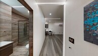 This thoughtfully designed single-floor space blends modern aesthetics with practical functionality. The open main area is anchored by continuous LED linear lighting recessed along the ceiling edges and ceiling plane, casting a clean, ambient glow across the gray wood-look flooring that runs throughout. A wall-mounted mini-split HVAC unit ensures year-round climate control, while dark curtain panels along one wall offer privacy and a refined, finished look.

The fully equipped kitchen features dark-toned cabinetry, white countertops, and a full suite of stainless steel and black appliances, including a gas range, microwave, refrigerator, and sink with a flexible faucet. A built-in desk area with a white countertop extends from the kitchen, creating a seamless workspace transition. A decorative textured wall panel adds an artistic accent to the kitchen corner.

The bathroom is a standout feature, finished with wood-look wall and floor tiles for a warm, spa-inspired atmosphere. It includes a floating vanity with a vessel sink, a backlit LED mirror, and a frameless glass-enclosed walk-in shower. The space is accessible, with grab bars thoughtfully incorporated near the toilet and shower.

Overall, the space presents a cohesive, contemporary design with high attention to detail, making it well-suited for use as a studio, office, or live-work environment.